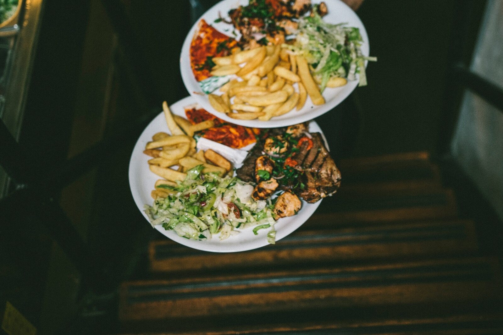 Twee borden met gegrild vlees, salade en friet worden de trap op gedragen